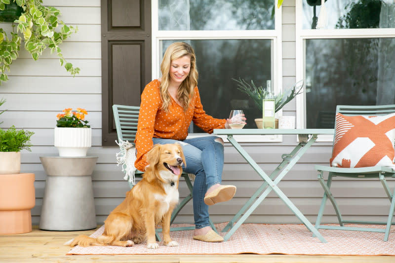 Blonde woman in orange shirt and jeans sits on a metal chair on the porch of a manufactured home that’s decorated with plants, holding a wineglass and petting a gold and white dog.
