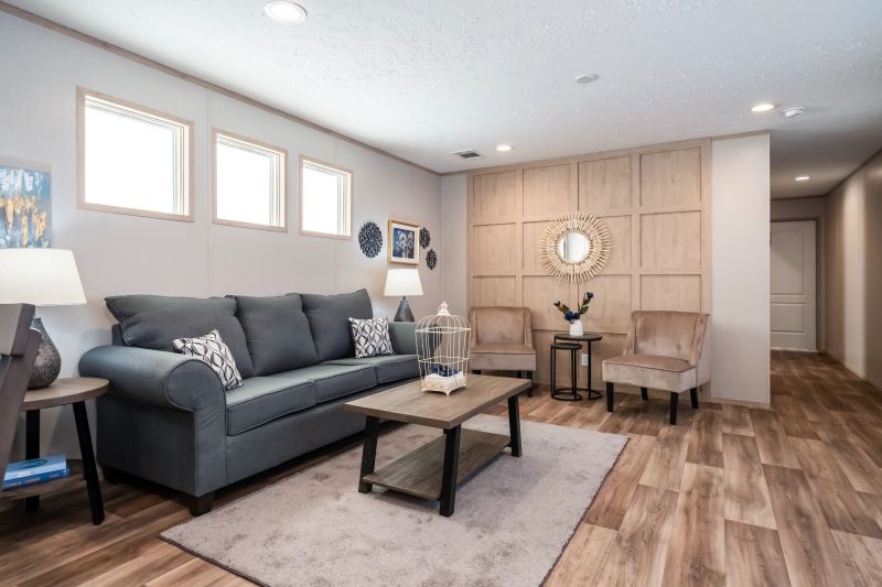 A manufactured home living room is in view with a light colored wood accent wall, three high cube mirrors over the dark grey couch and  other neutral decor.