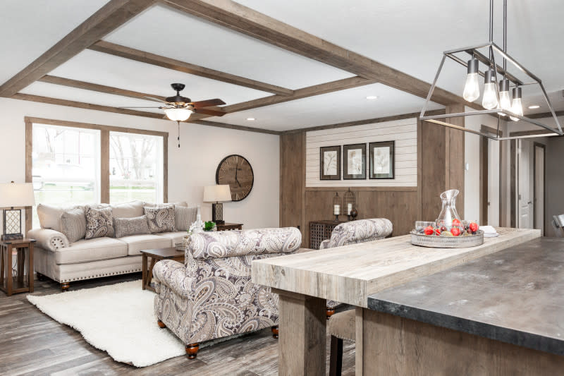 Manufactured home living room with shiplap accent wall, ceiling beams and neutral decor, with kitchen island and rectangular metal chandelier in the foreground.