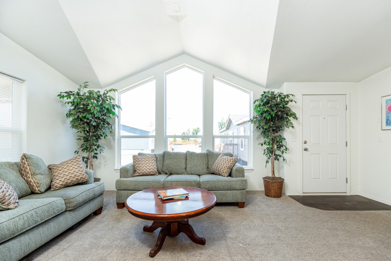 Floor to ceiling windows come to a point, letting light fill the room in this manufactured home living room. There are two fake trees for decoration on either side of the window, two light green couches, and a cherry wood colored coffee table.