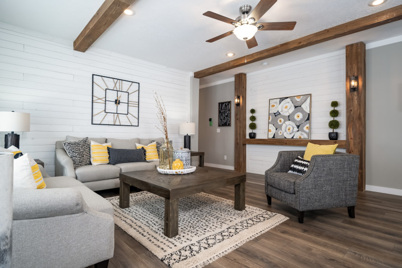 A manufactured living room is in view with a white ship lap wall, wooden beam on the ceiling, and a focus wall. There’s gray furniture and a wooden coffee table.