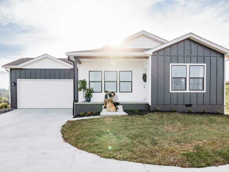 A couple sits on the front porch steps of their gray-sided home while petting their dog.