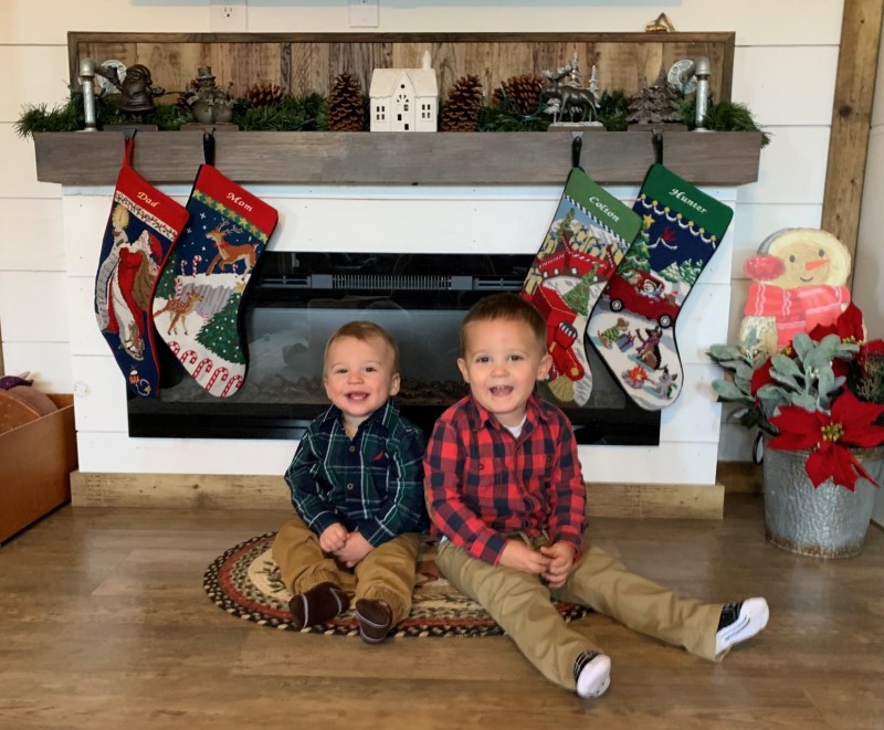 Two young boys in plaid shirts sitting on the floor in front of a fireplace.