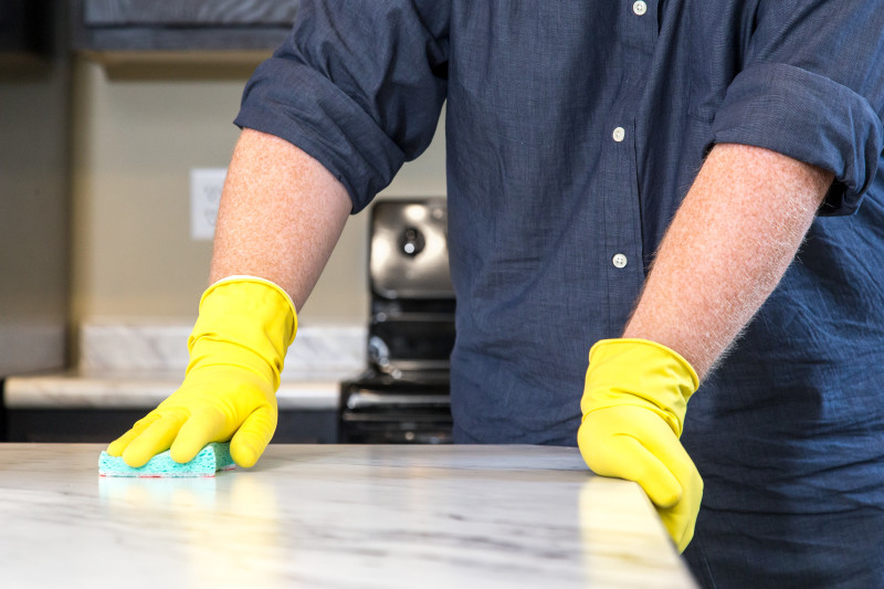 A man in a blue shirt with yellow rubber gloves uses a light blue sponge to clean a marble countertop in a manufactured home kitchen.