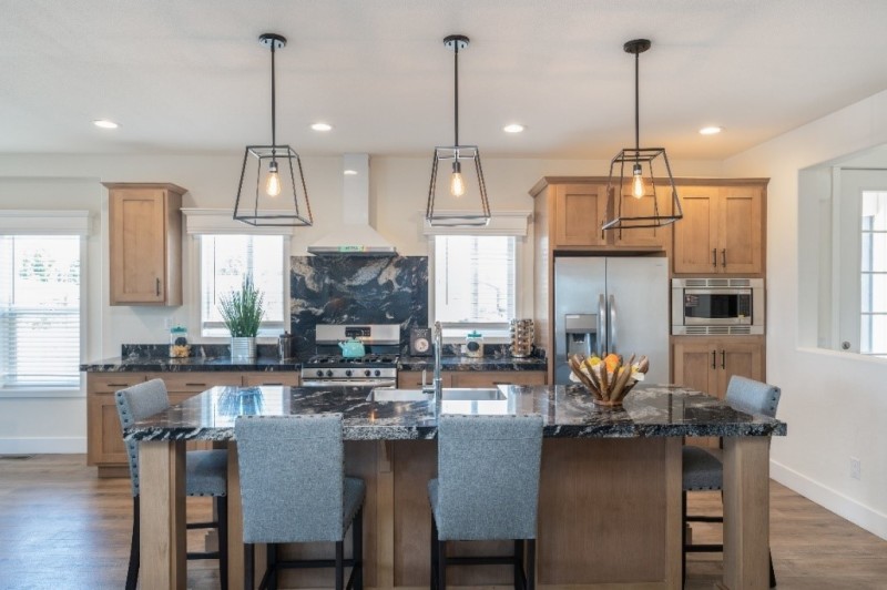 Classic kitchen in the Coronado manufactured home with honey-colored cabinets, white walls and marble-style countertops. Appliances are stainless steel and it has a large kitchen island in the center with chairs around it. 