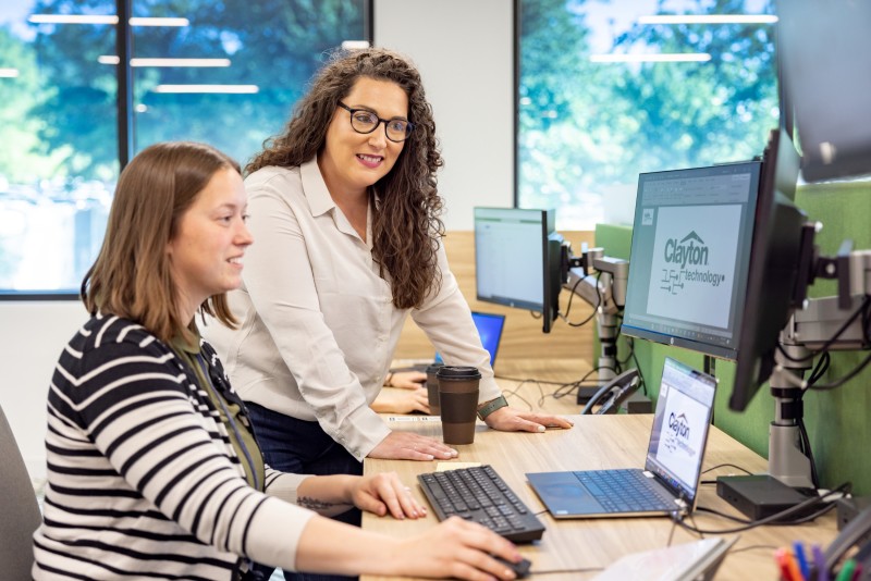 Woman stands behind another seated woman to view a computer monitor screen, with more office equipment and windows in the background.