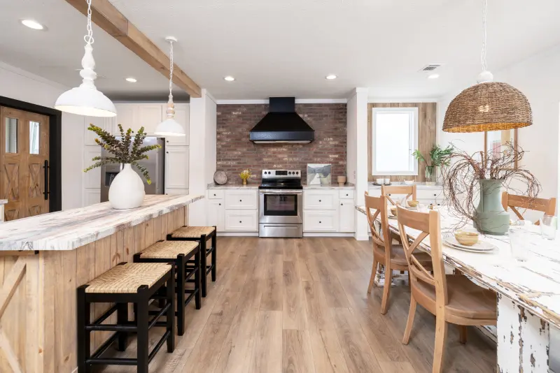 A manufactured home kitchen with a rustic island, brick backsplash behind a black and stainless-steel stove and range hood, white cabinets, and a long dining table.