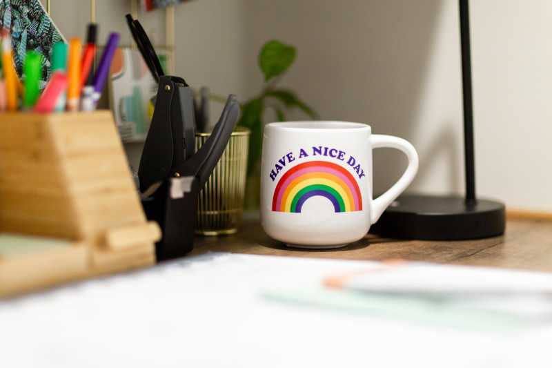 A closeup of an office desk that focuses on a white coffee cup that reads “Have a Nice Day” above a rainbow, a container of markers and a hole punch beside it.