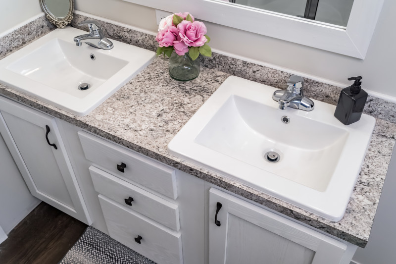 Manufactured home bathroom with dark wood floor, black, white and pink granite style countertops, white cabinets, sinks, walls and mirror and silver faucets, with a vase of pink flowers on the counter