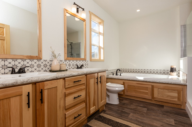 A side angle of a primary bathroom shows a double sink vanity with light wood cabinets, a playful, starburst backsplash, and a soaker tub and toilet to the side.