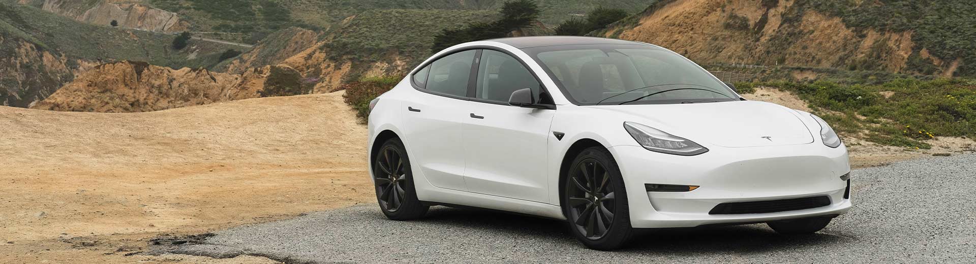 En-vit-Tesla-parkerad-på-en-grusväg-framför-dimmiga,-gröna-kullar-och-ett-bergigt-landskap