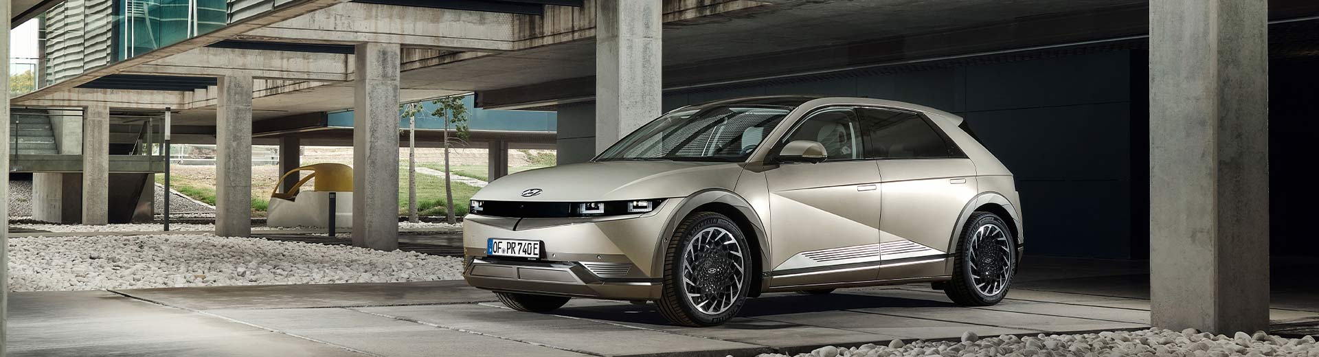 En-champagnefärgad-Hyundai-Ioniq-5-parkerad-under-en-modern-betongstruktur-med-lameller.-Bilen-visas-snett-framifrån