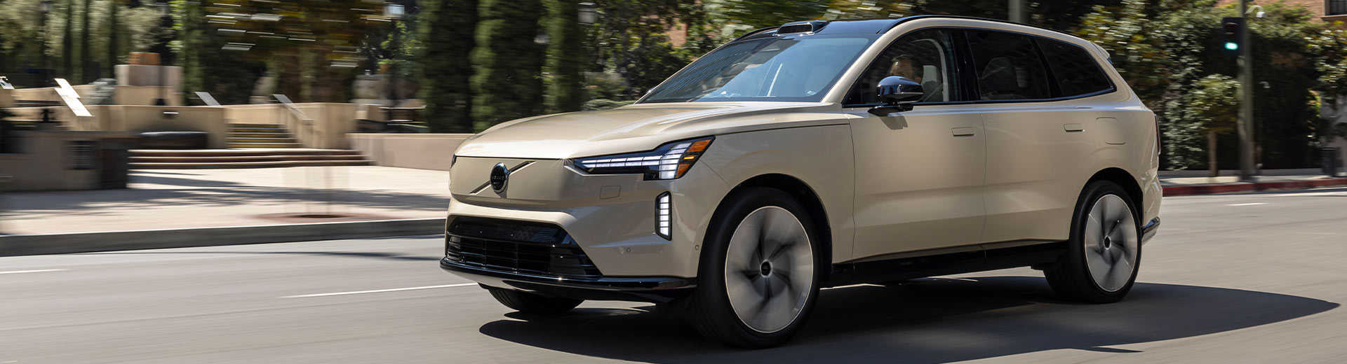 En-sandfärgad-Volvo-EX90,-en-helelektrisk-sjusitsig-SUV,-körs-genom-en-stadsmiljö-med-höghus-i-bakgrunden