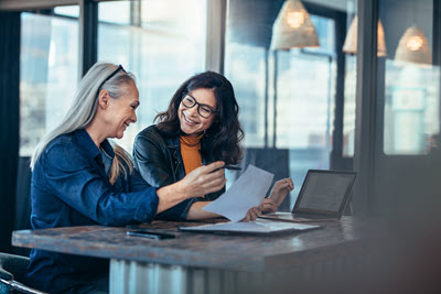 Stockfoto: Zwei Frauen lachend am Tisch mit Papieren und Laptop