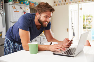 Mann mit Laptop, Handy und Kaffeebecher am Küchentisch