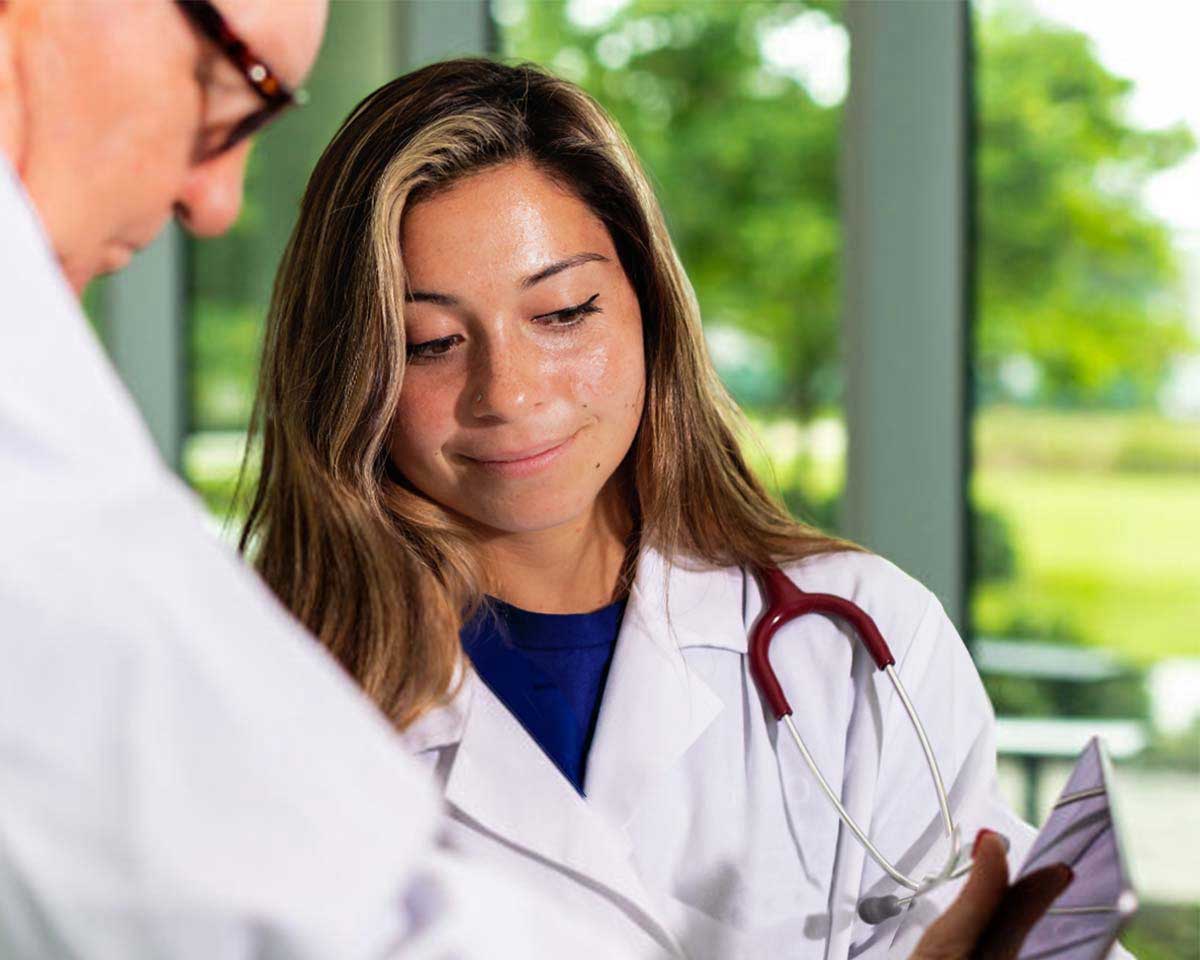 Lynn Healthcare student and instructor in lab coats viewing a tablet in front of windows overlooking a manicured landscape