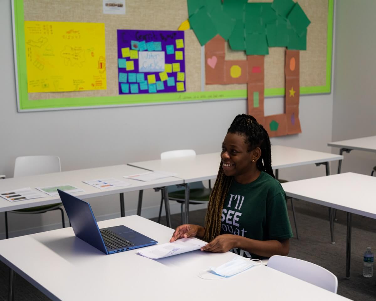 Elementary education student smiling, sitting at a classroom desk in front of her laptop and sorting paperwork
