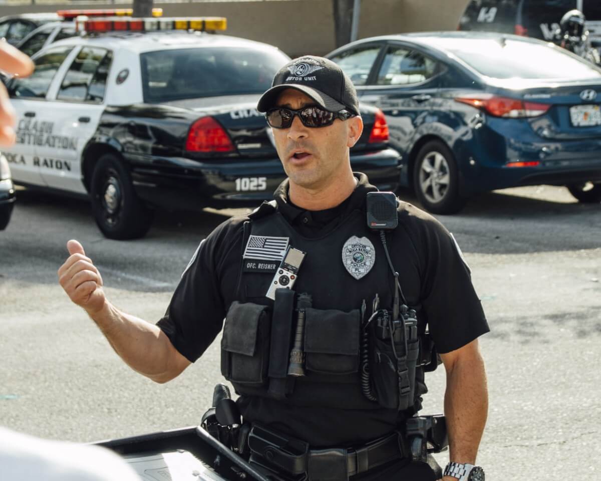 Police officer in bullet-proof vest standing outside and talking to students with a law enforcement vehicle in the background