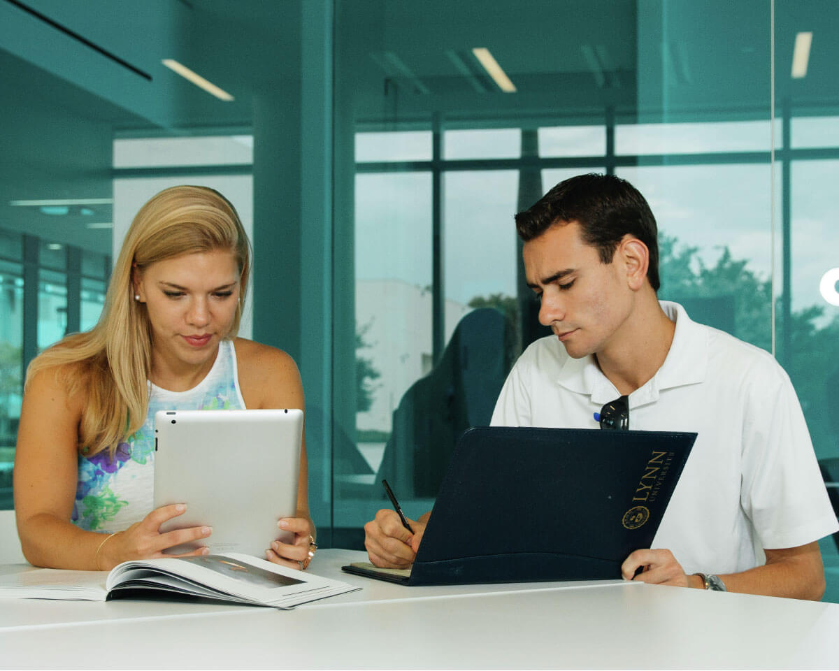 Two investment management students working together with tablet and laptop in modern study room with large windows