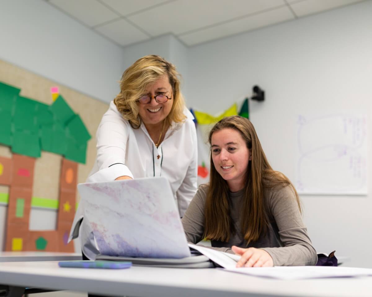 Elementary education student working on a laptop in a classroom with her instructor smiling and pointing to something on the laptop screen