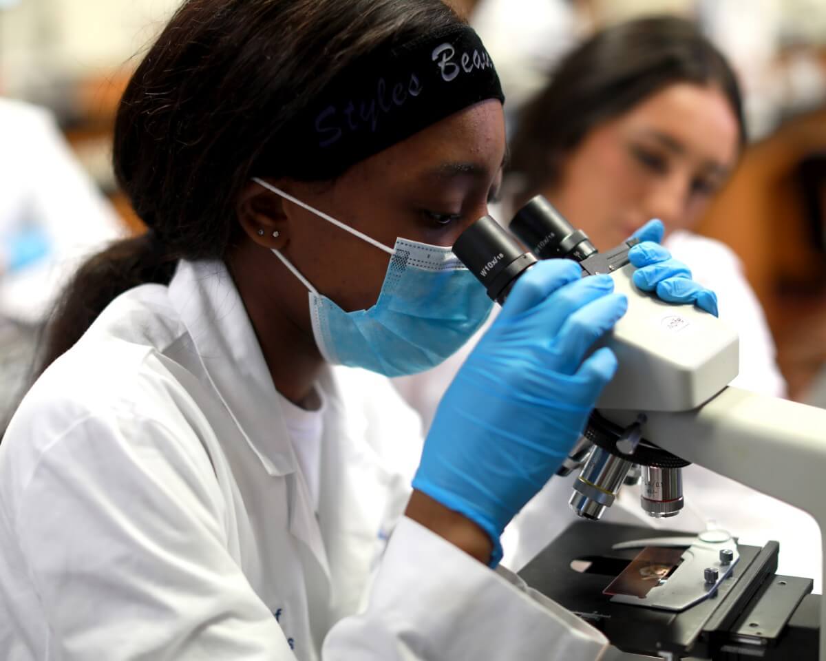 Lab technician wearing labcoat, mask, and gloves looking at a specimen through a microscope