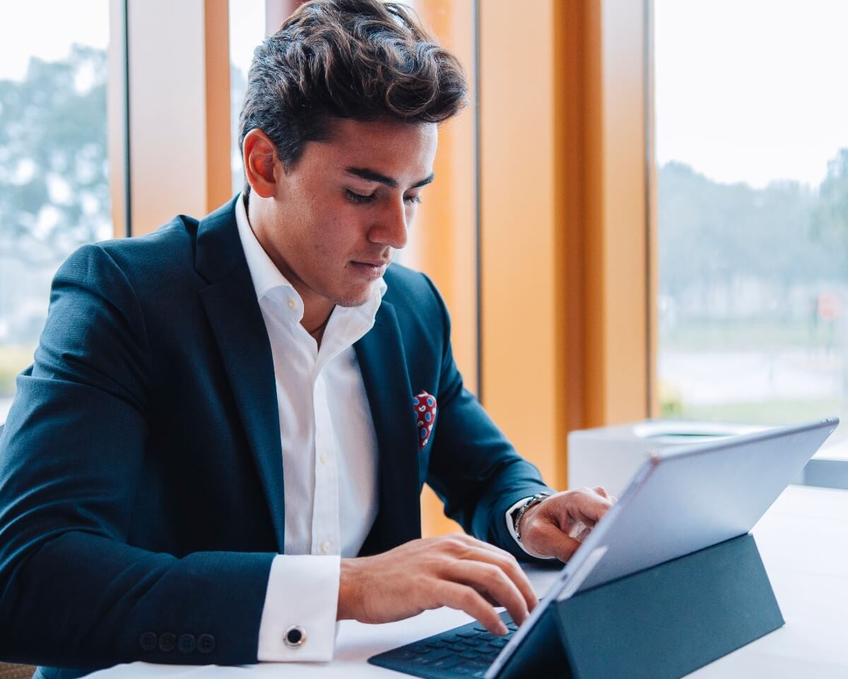 Lynn student in a suit working on laptop at a table in front of windows overlooking a tree-lined park