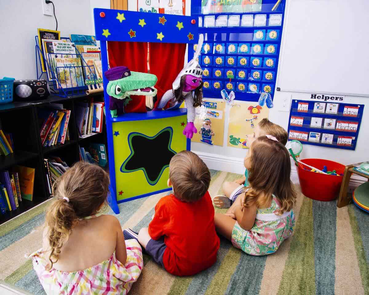 Four young children sitting cross-legged on the floor of their classroom, watching an educational puppet show