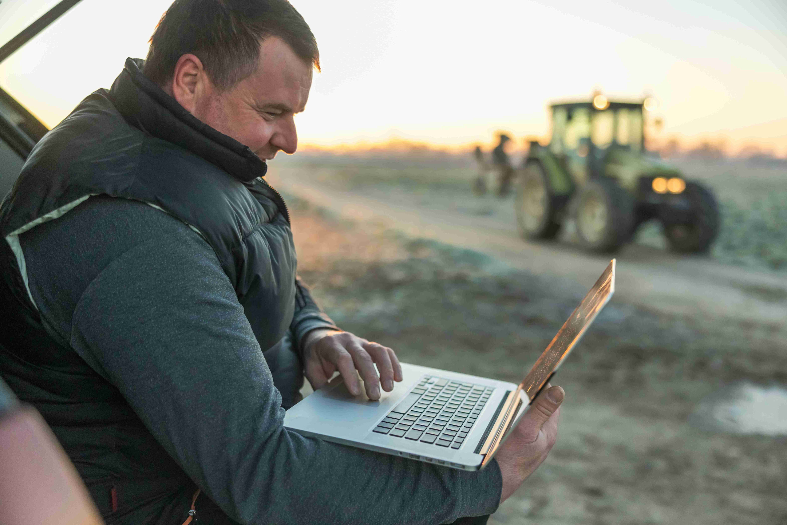 A man working on a laptop while seated on a dirt road, likely managing farm payroll tasks.
