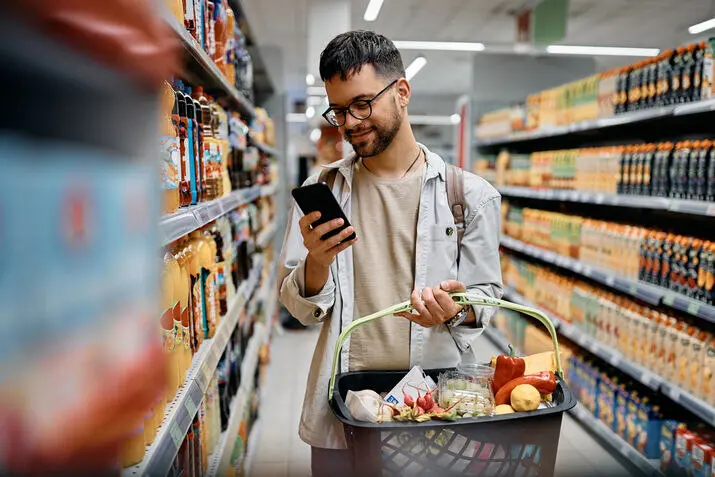 A man using his smartphone in a supermarket, illustrating modern retail merchandising practices.