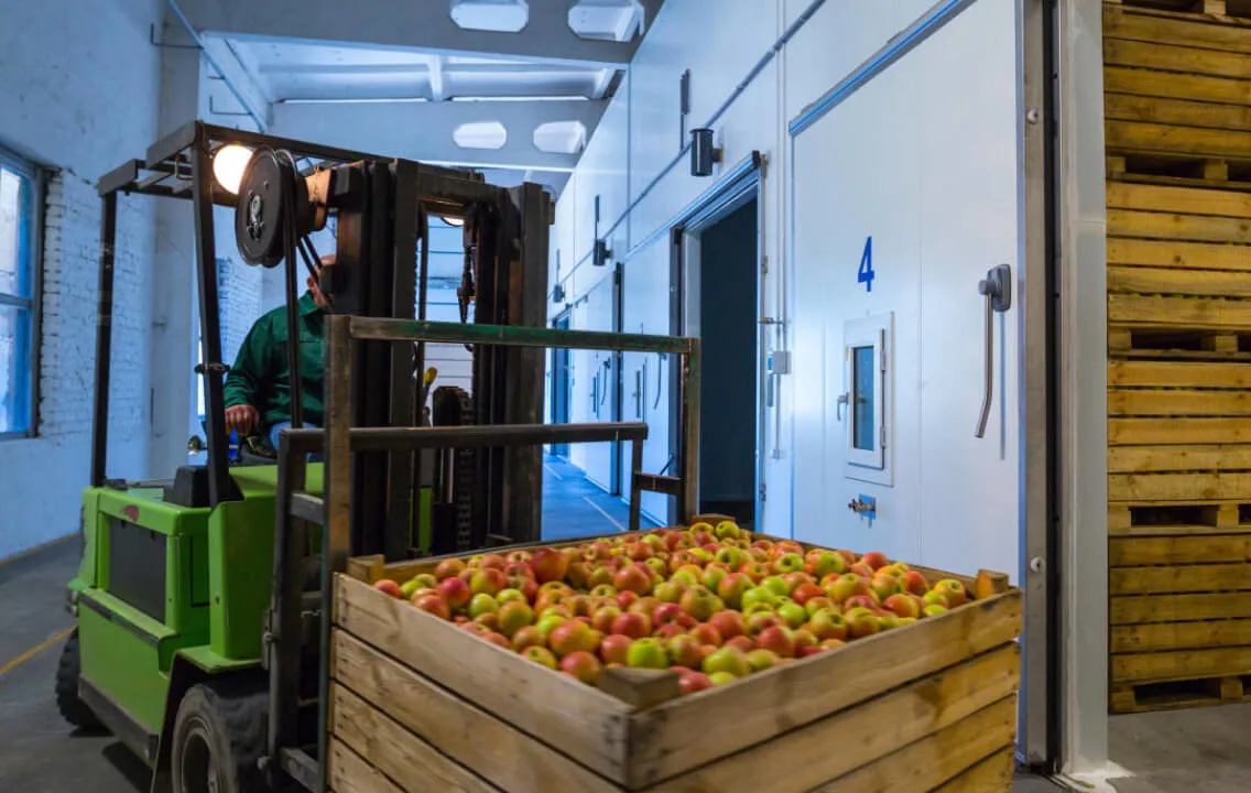 A man operating a forklift is moving a crate of apples