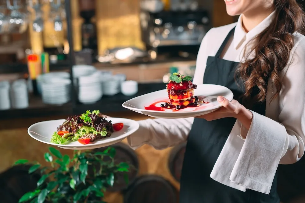 A waiter with a linen napkin draped over their arm is carrying two plates to a table: one with a field greens salad, and the other with a fancy plating of sliced fruit stacked and topped with a blueberry reduction and sprig of mint.