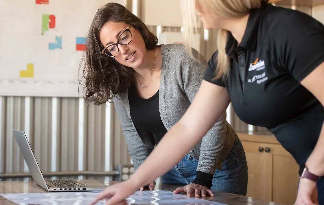 Two women with a laptop standing around a table looking at maps