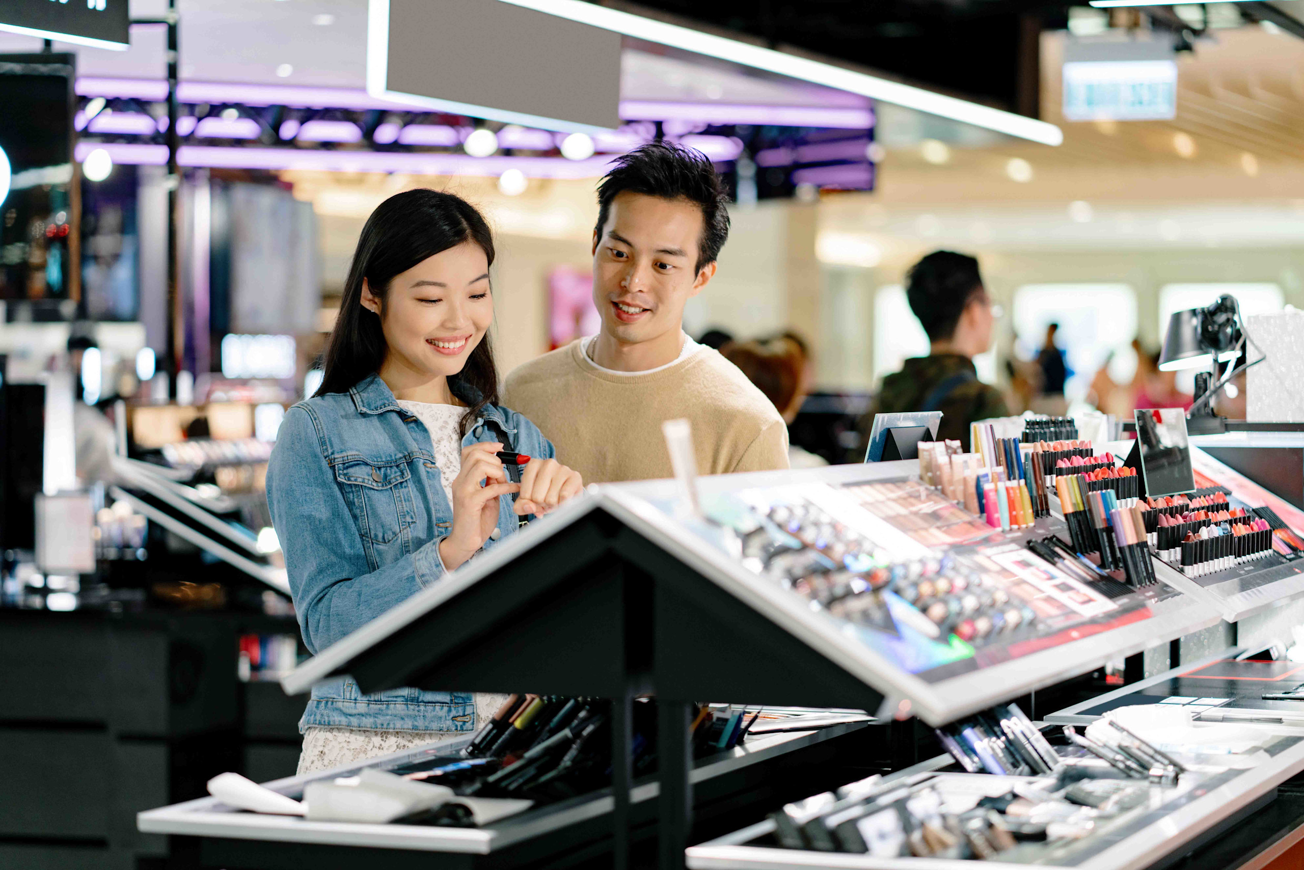 A man and woman stand at a cosmetics counter in a travel retail setting, exploring beauty products together.