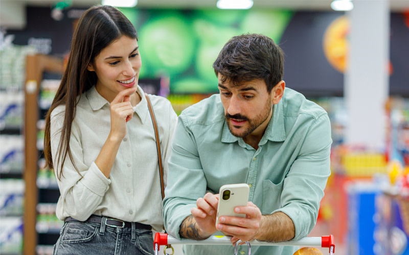 A couple standing in a grocery store aisle looking at a smartphone together while shopping with a cart.