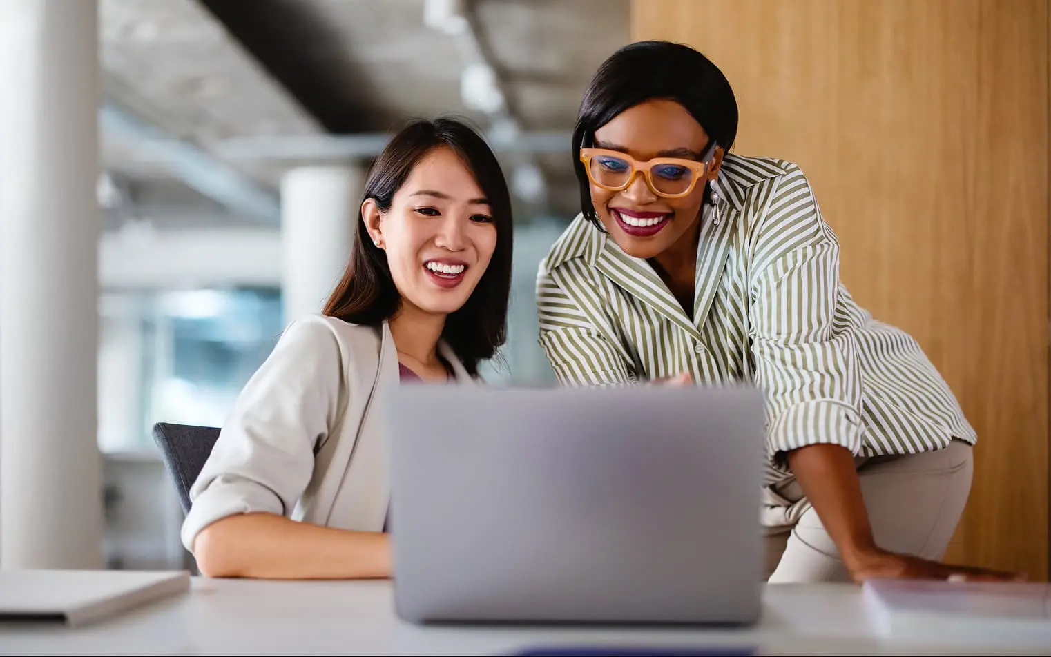 Two professional women collaborating at a laptop in a modern office, smiling while reviewing content together