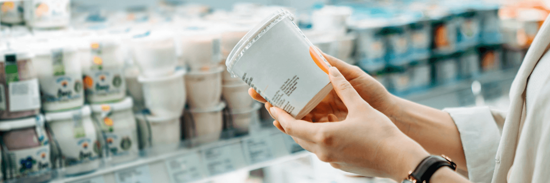 A person holding a yogurt package in front of grocery store shelves filled with various dairy products.