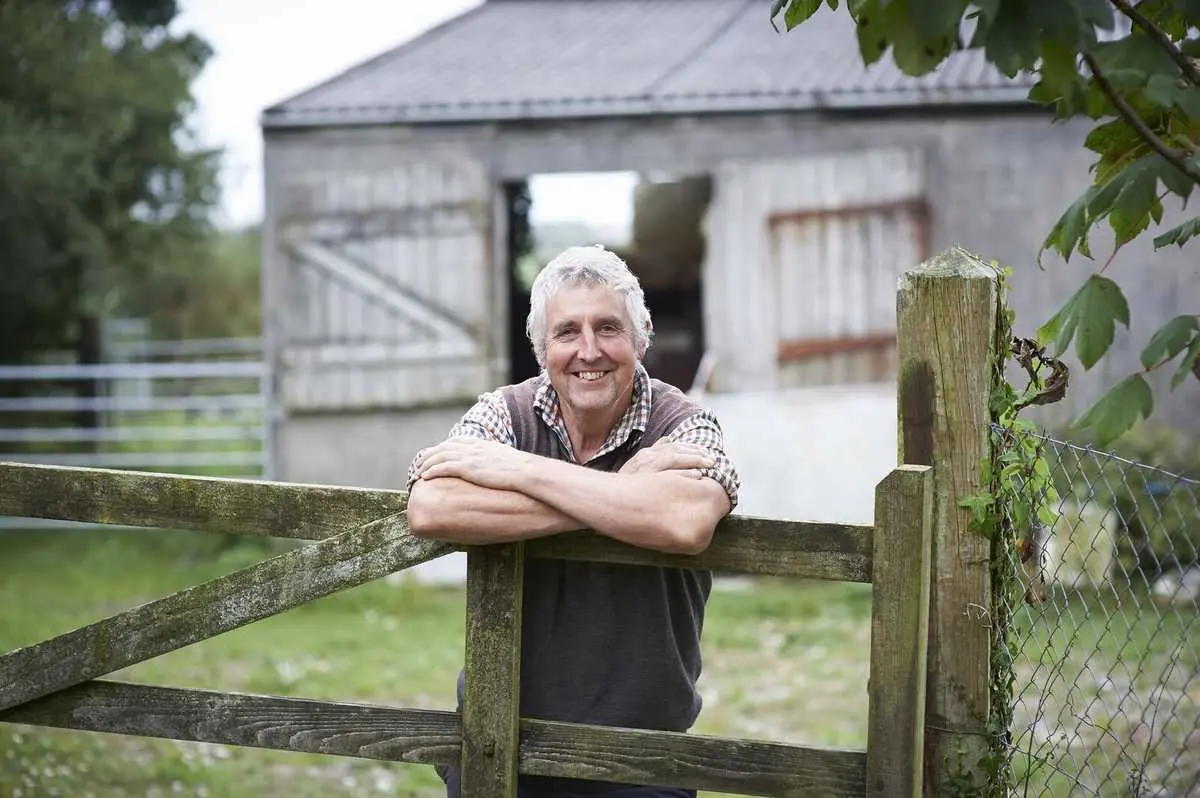 Smiling older male farmer wearing a plaid shirt and vest, leaning on a wooden gate in front of a barn.