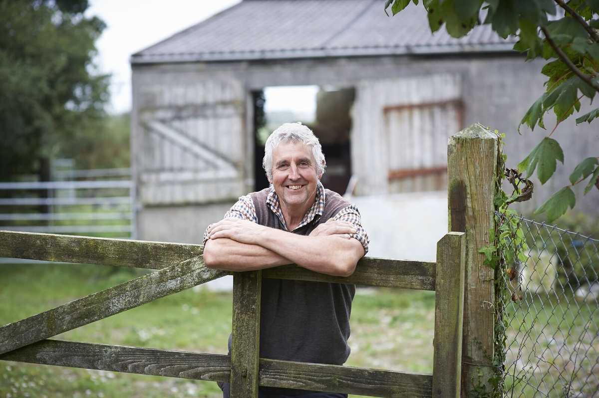 Smiling older male farmer wearing a plaid shirt and vest, leaning on a wooden gate in front of a barn.