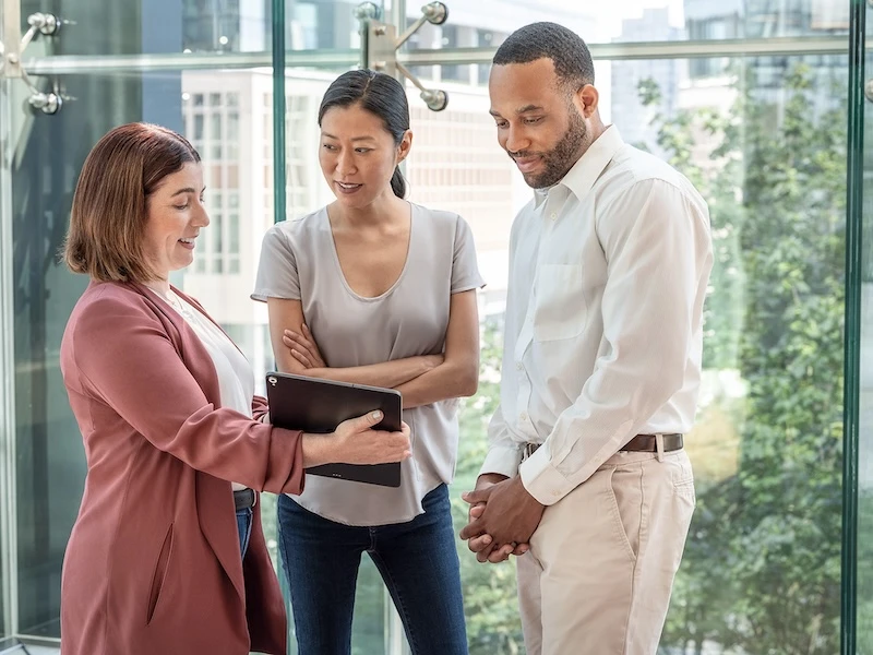 Three casually dressed professionals are gathered by a window, one of whom is presenting information to the other two on a tablet.