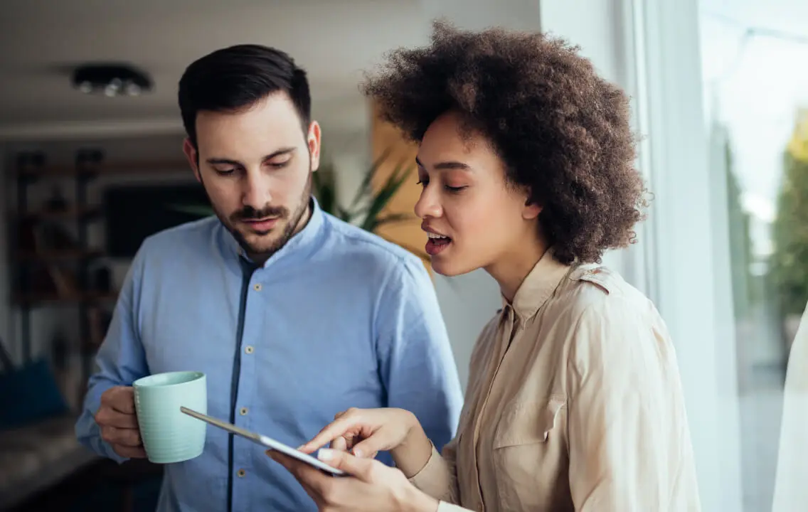 A man and woman looking at a tablet while standing in front of a window.