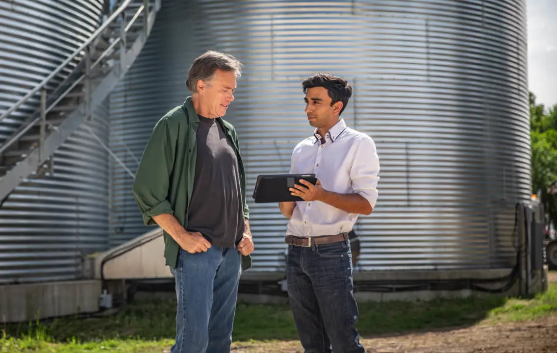 Two men standing in front of a metal silo, looking at a tablet