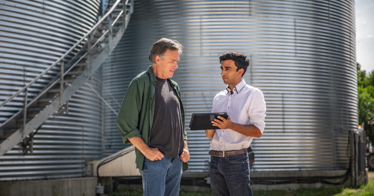 Farmer and advisor in front of silos, looking at tablet.