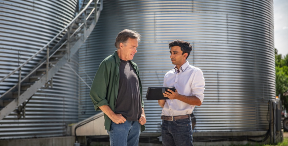 Farmer and advisor in front of silos, looking at tablet.