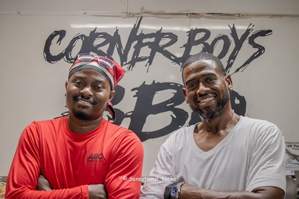Two men smiling standing in front of a sign for their restaurant