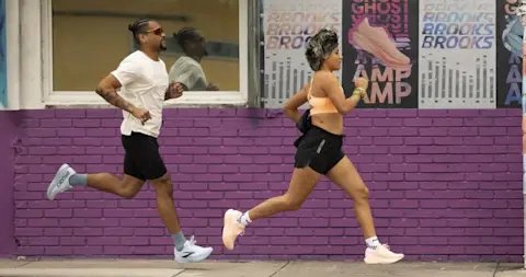 Runners along sidewalk with purple brick background running in the Brooks Ghost Amp running shoes