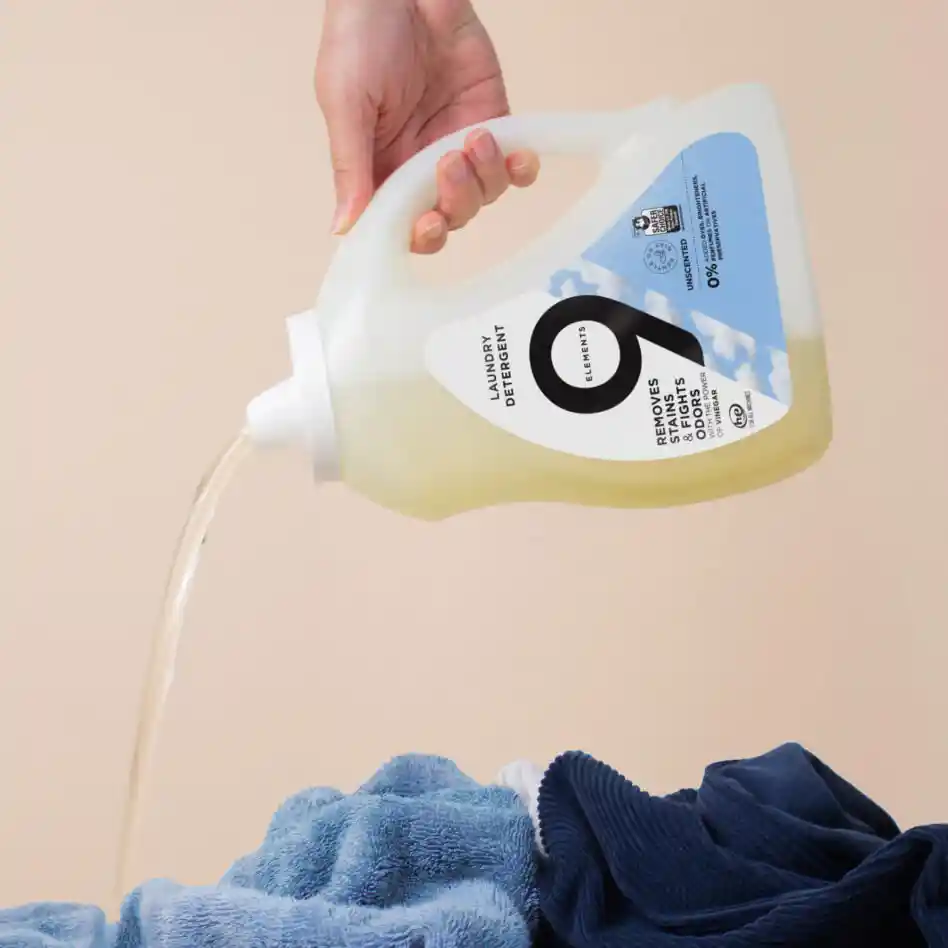 A hand holding 9 Elements unscented laundry detergent bottle and pouring it on the clothes