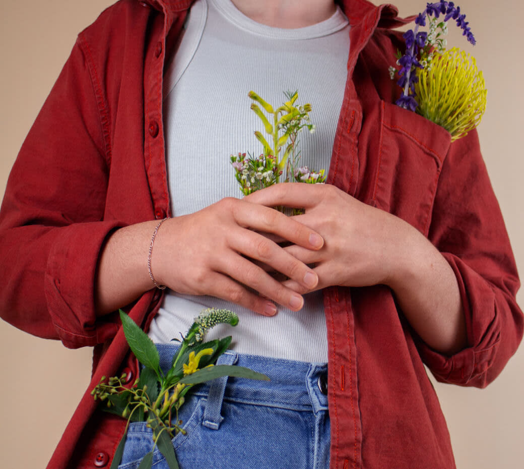 Central part of a woman's body dressed in blue jeans, red jacket and white shirt with pockets filled with live plants and flowers.