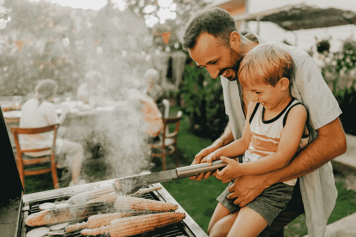 Vater grillt mit seinem Sohn im Garten und die Nachbarn sind zu Gast.