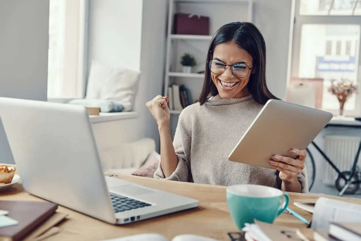 Eine lächelnde Frau mit Brille blickt auf ihr Tablet und feiert einen finanziellen Erfolg, was symbolisiert, wie man am besten sparen kann.