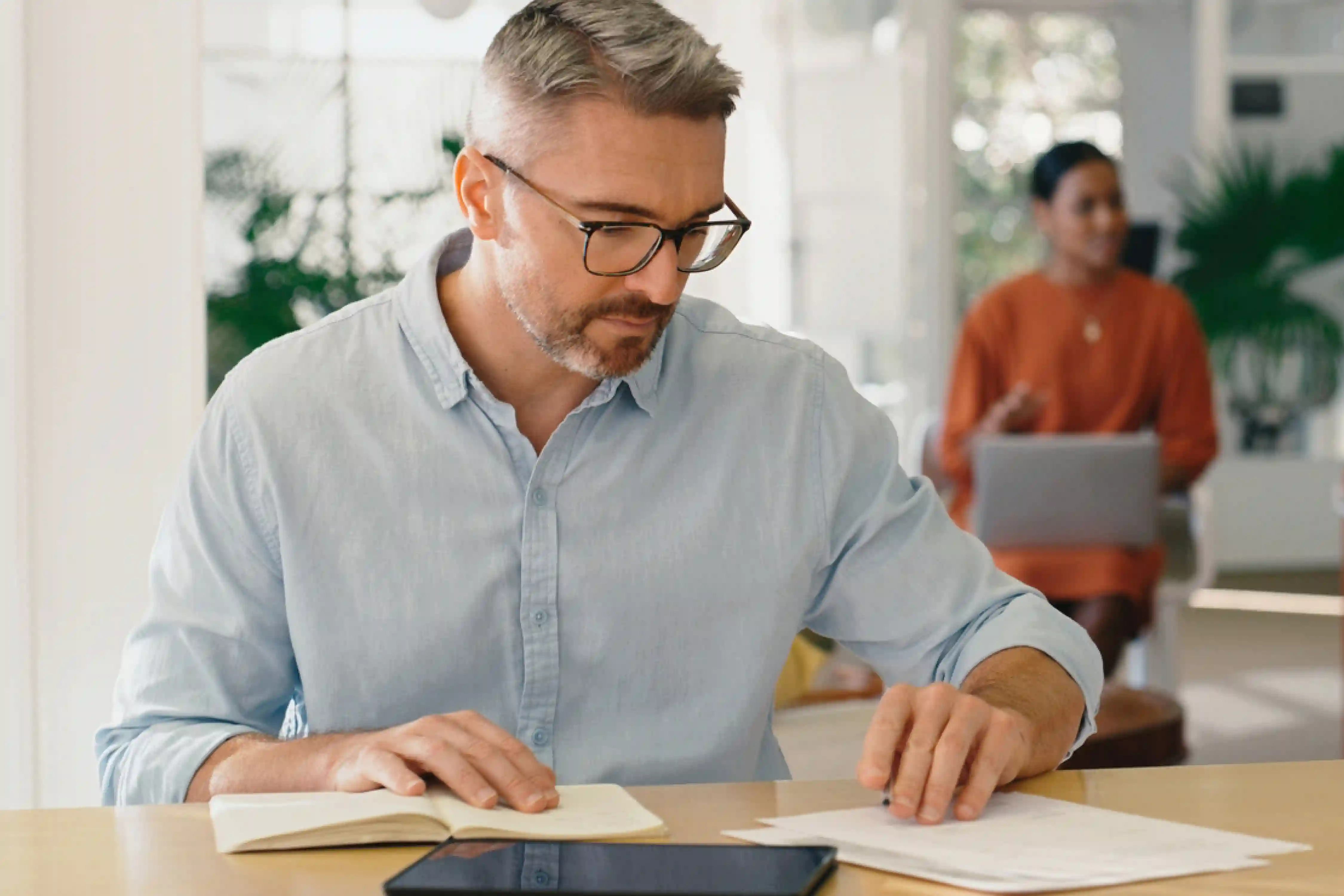 A middle-aged man fills out applications for the granting of subsidies for an energy-efficient renovation. His wife is in the background looking after an advisor.